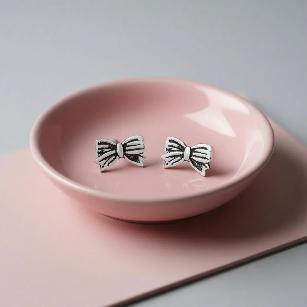 A pair of silver earrings with black bow designs are placed on a pink ceramic dish.